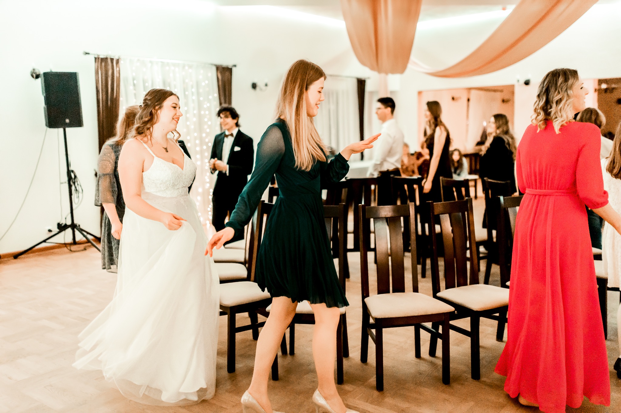 Panna młoda w koronkowej sukni ślubnej i goście w sukienkach wieczorowych w sali bankietowej z krzesłami ustawionymi w rzędach.