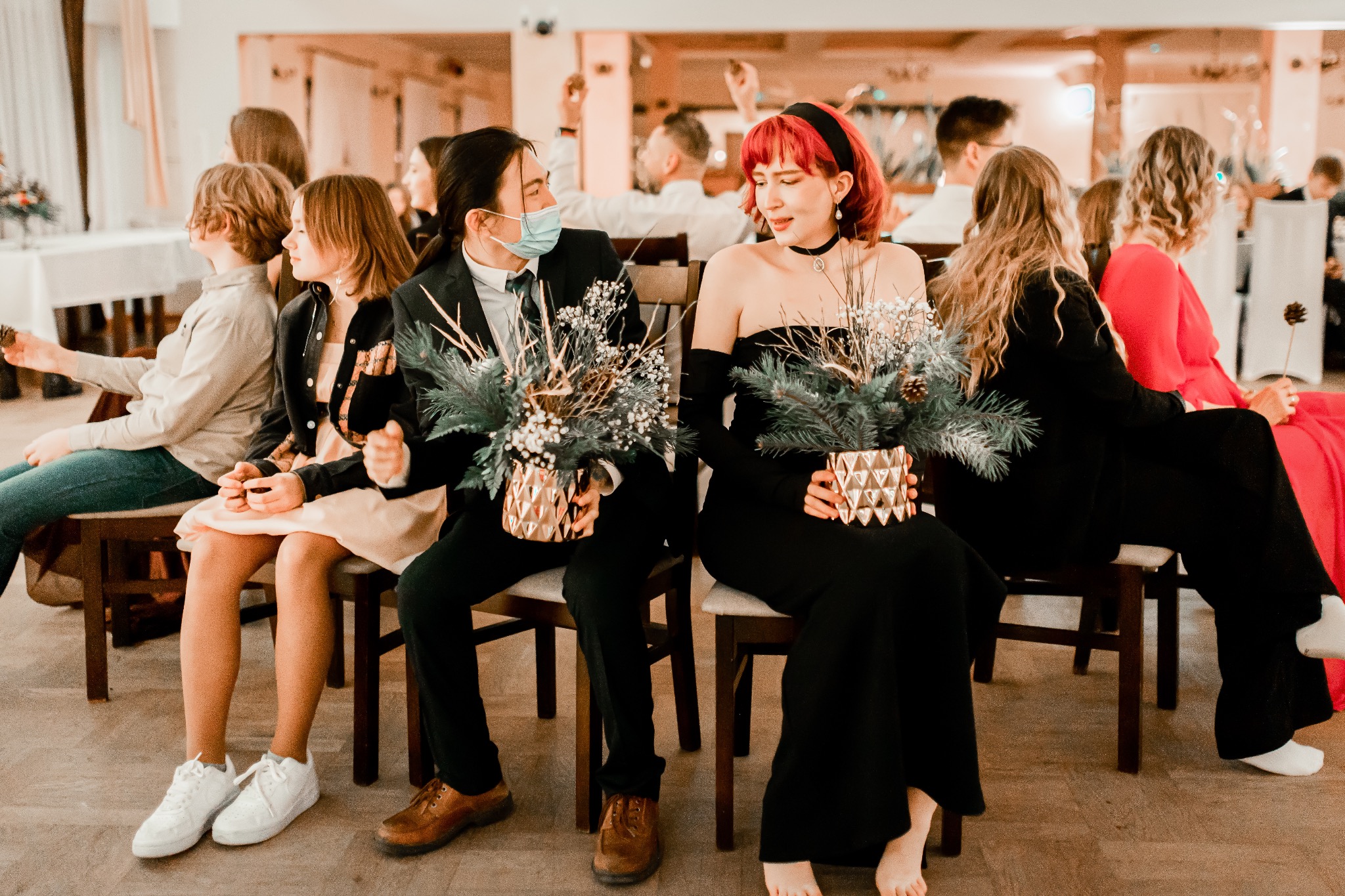Goście weselni siedzą na krzesłach i trzymają złote wazony z kompozycjami kwiatowymi z igliwiem, mężczyzna w garniturze i maseczce, kobieta w czarnej sukni bez ramiączek i z czerwoną czupryną.