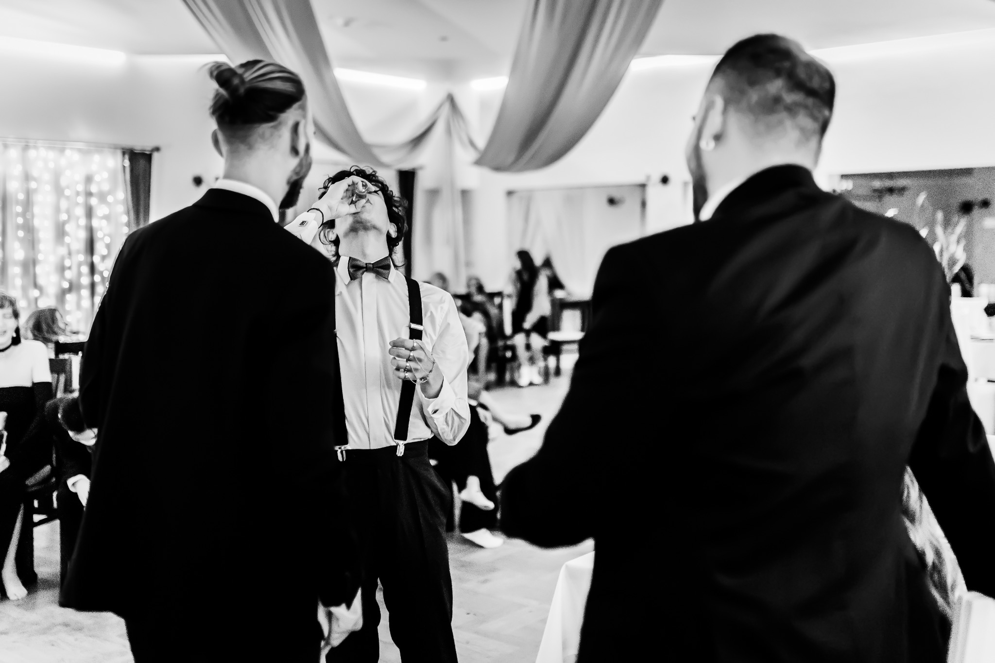 Mężczyzna w muszce i szelkach wznosi toast podczas uroczystości, widziany z perspektywy dwóch osób w garniturach, tło z dekoracjami.