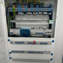 Otwarta skrzynka elektryczna z widocznymi bezpiecznikami, przewodami w różnych kolorach i elementami montażowymi, zainstalowana na białej ścianie.