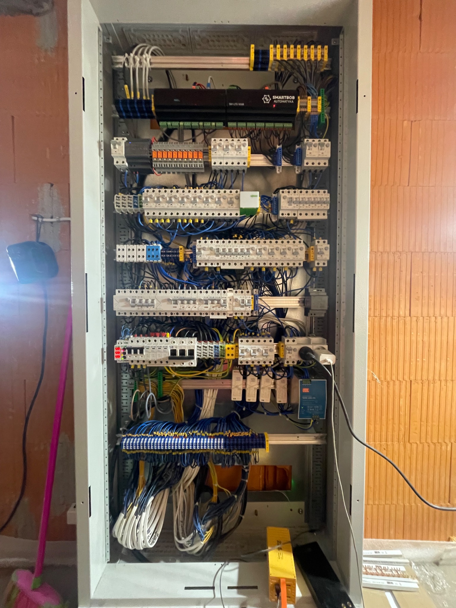 Otwarta szafa sterownicza z widocznymi elementami automatyki, okablowaniem i zabezpieczeniami, umieszczona we wnętrzu z surowymi ścianami z cegły.