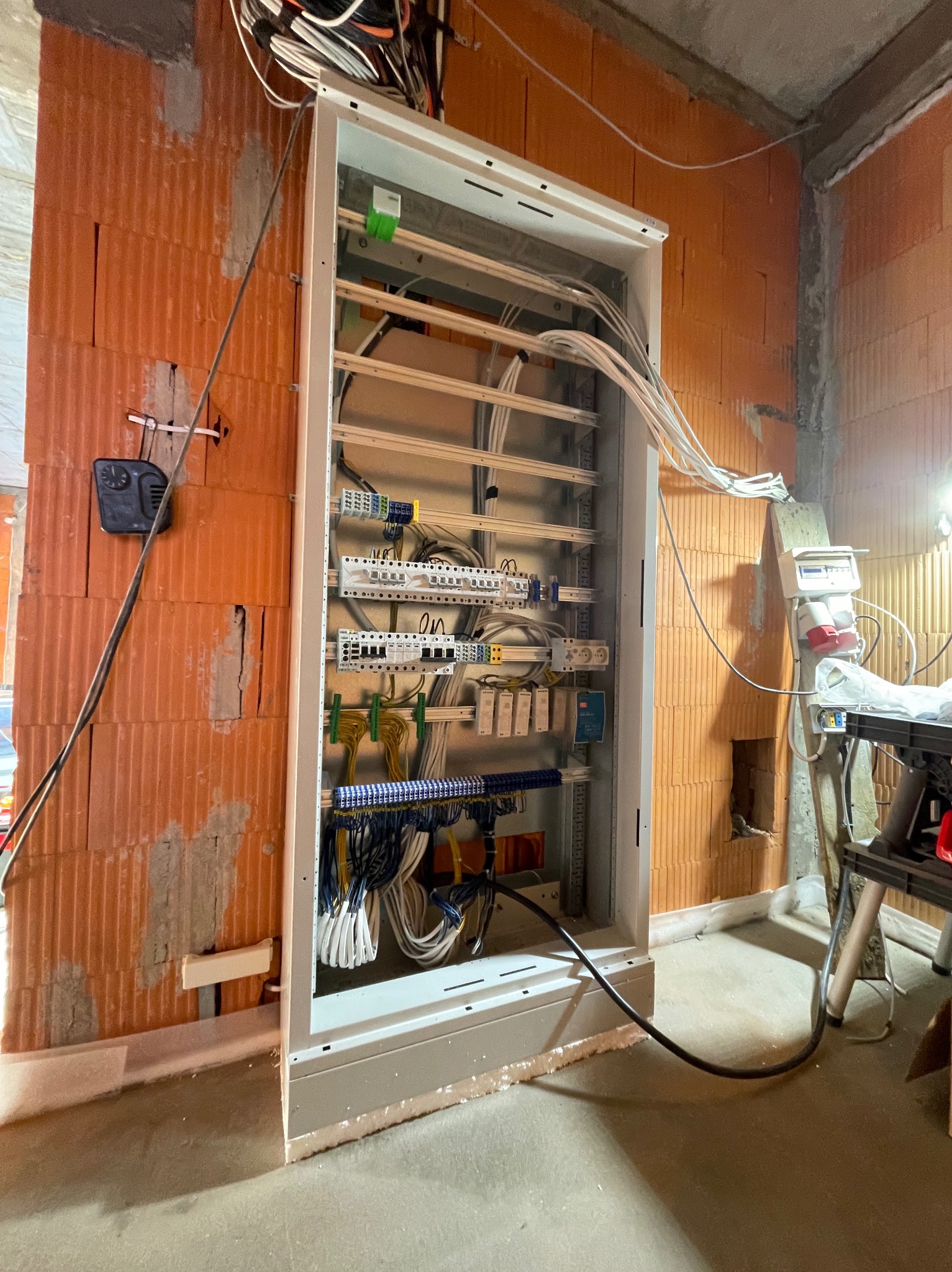 Otwarta skrzynka rozdzielcza z podłączonymi przewodami w surowym wnętrzu budynku, widoczne ceglane ściany i fragmenty instalacji elektrycznej.