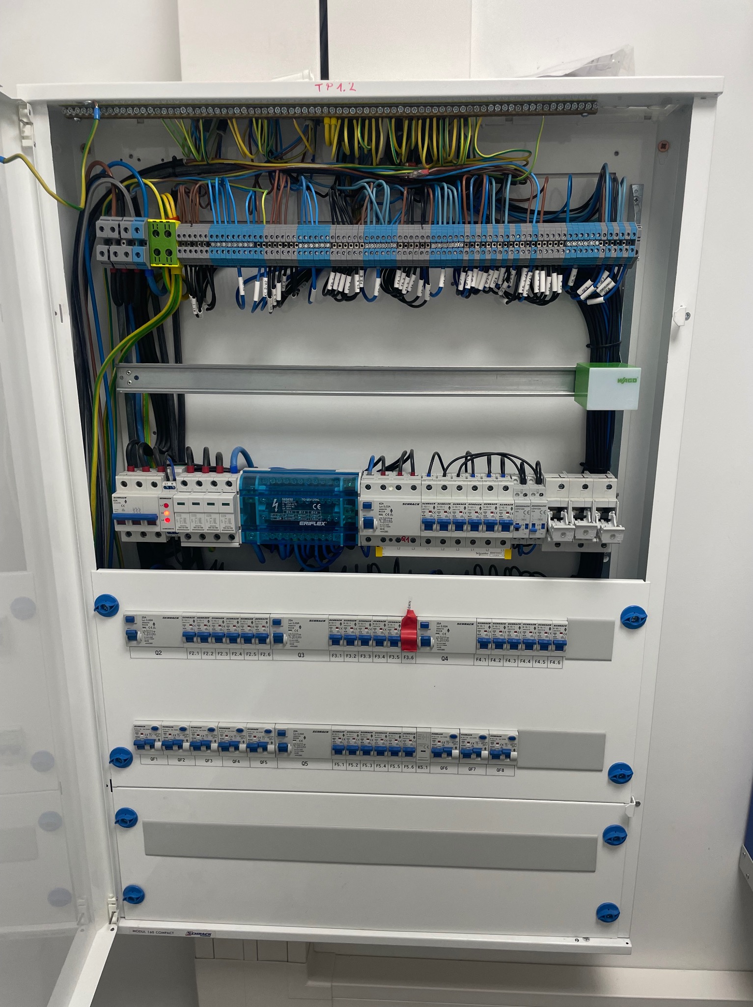 Otwarta skrzynka elektryczna z widocznymi bezpiecznikami, przewodami w różnych kolorach i elementami montażowymi, zainstalowana na białej ścianie.