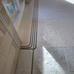 ElektroSan Paweł Sanik - Wnętrze w trakcie prac wykończeniowych: widok z góry na trzy szare peszle elektryczne prowadzone po podłodze wzdłuż ściany z oknem, z fragmentem stopy w czarnym bucie w prawym dolnym rogu.