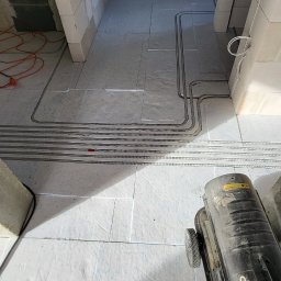 ElektroSan Paweł Sanik - Rury instalacyjne ułożone w bruzdach w podłodze w surowym wnętrzu budynku, widoczny nagrzewnica powietrza i fragment buta.