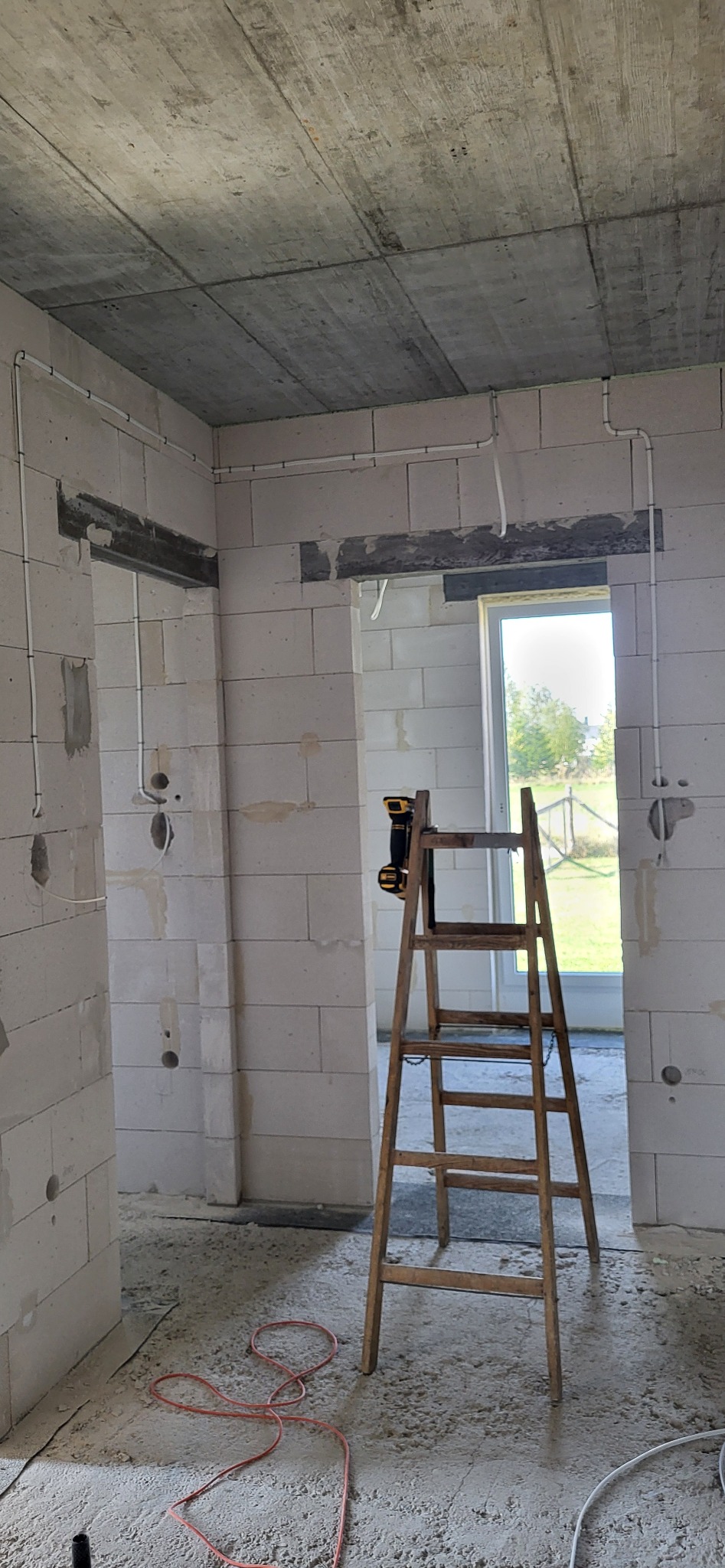 Wnętrze w trakcie prac wykończeniowych, widoczne kable elektryczne poprowadzone po ścianach z bloczków, drewniana drabina i zwój czerwonego kabla na podłodze.