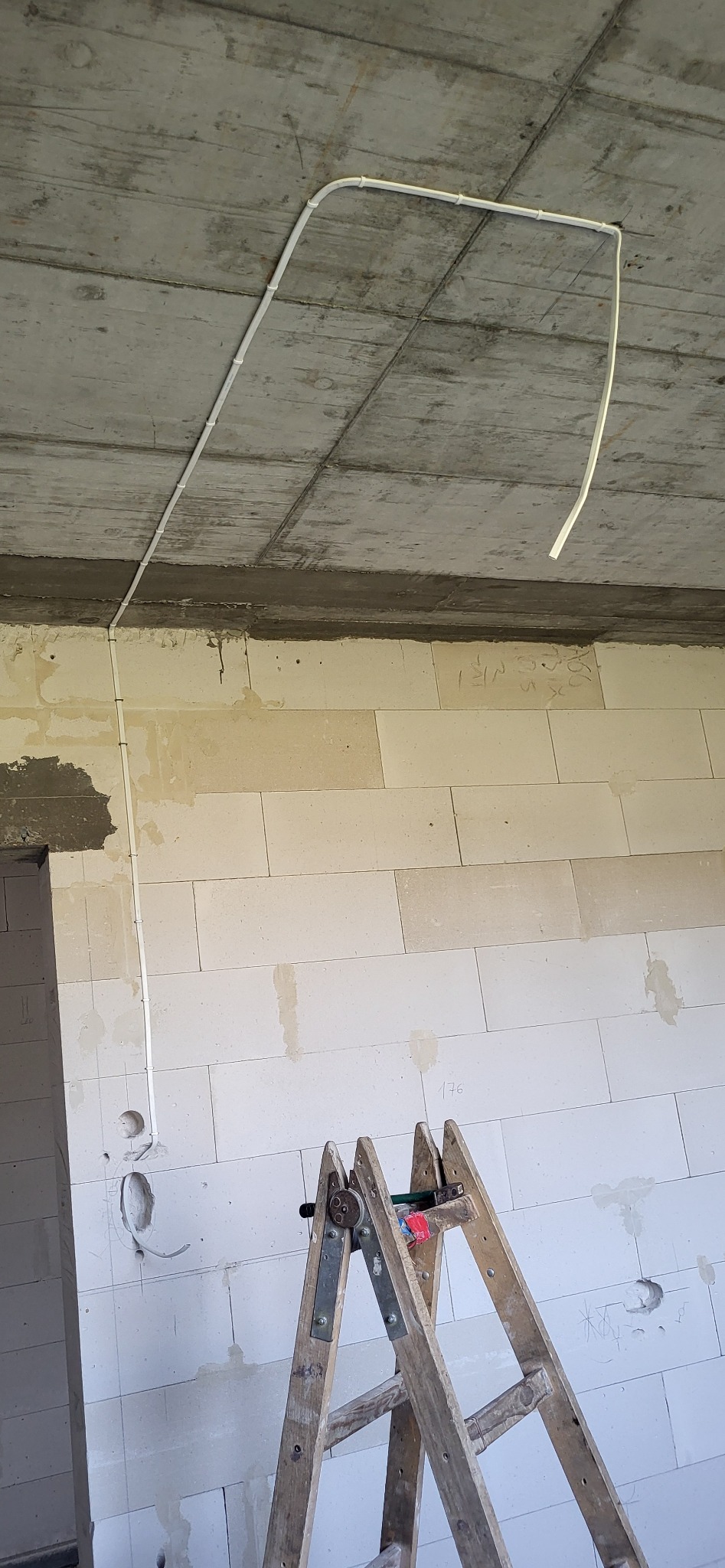 Wnętrze w trakcie prac wykończeniowych: widoczne kable elektryczne poprowadzone po suficie i ścianie z białego bloczka, drewniana drabina oparta o ścianę, surowy betonowy strop.
