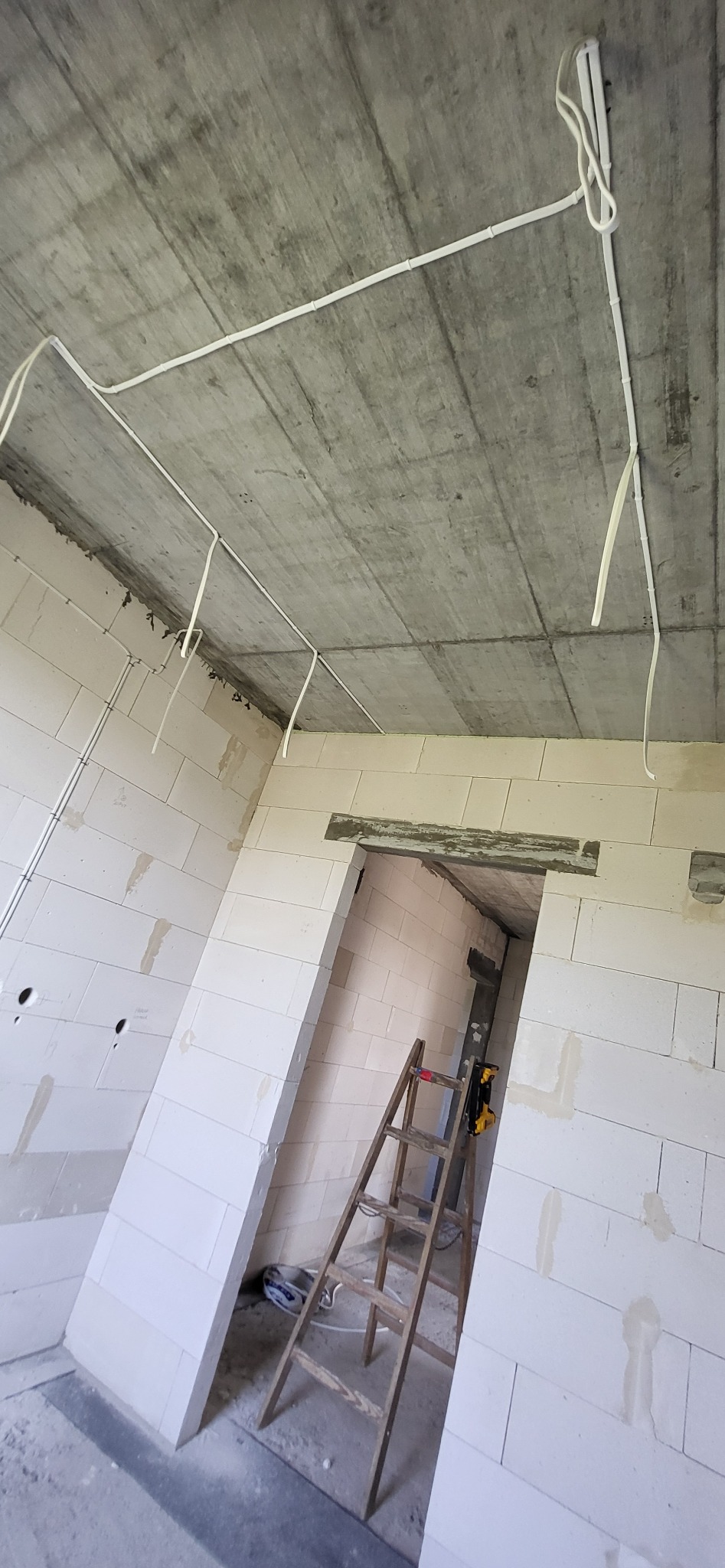 Surowy betonowy sufit z ułożonymi białymi kablami elektrycznymi w nowym budynku z białych bloczków, drewniana drabina oparta o ścianę w otworze drzwiowym.