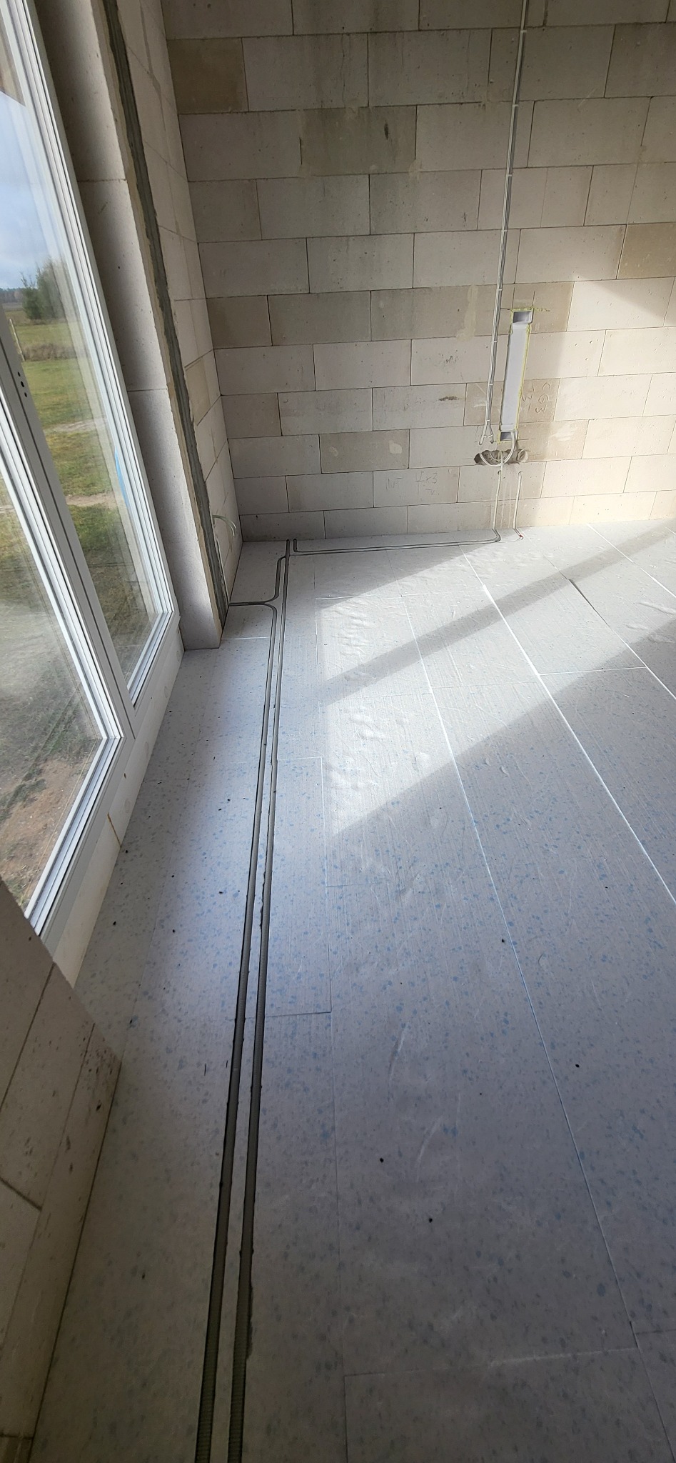 Wewnętrzne ściany z bloczków i podłoga w surowym stanie z widocznymi bruzdami pod instalacje elektryczne, rury prowadzone po ścianie pod sufitem.