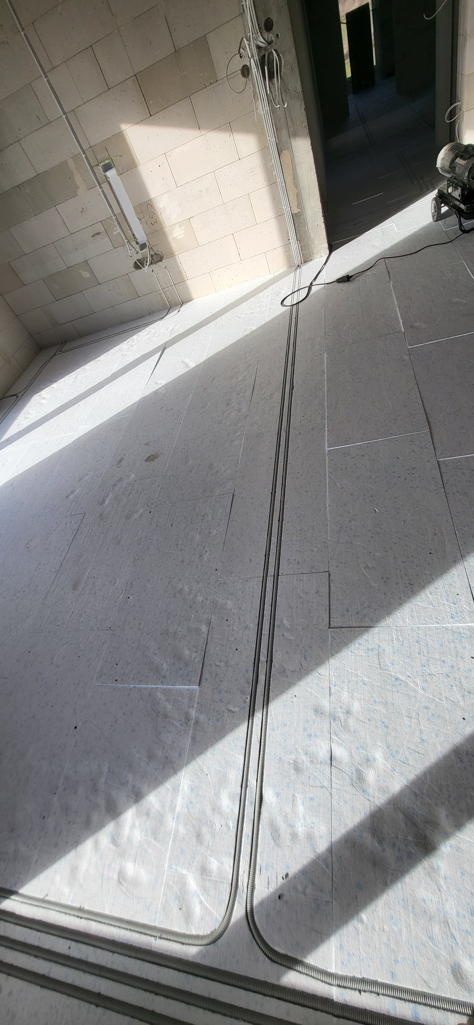 Surowy stan budynku z widocznymi przewodami elektrycznymi ułożonymi w peszlach na podłodze, prowadzącymi do ściany z pustaków, z widocznym oświetleniem technicznym na suficie i przewodami w korytkach.