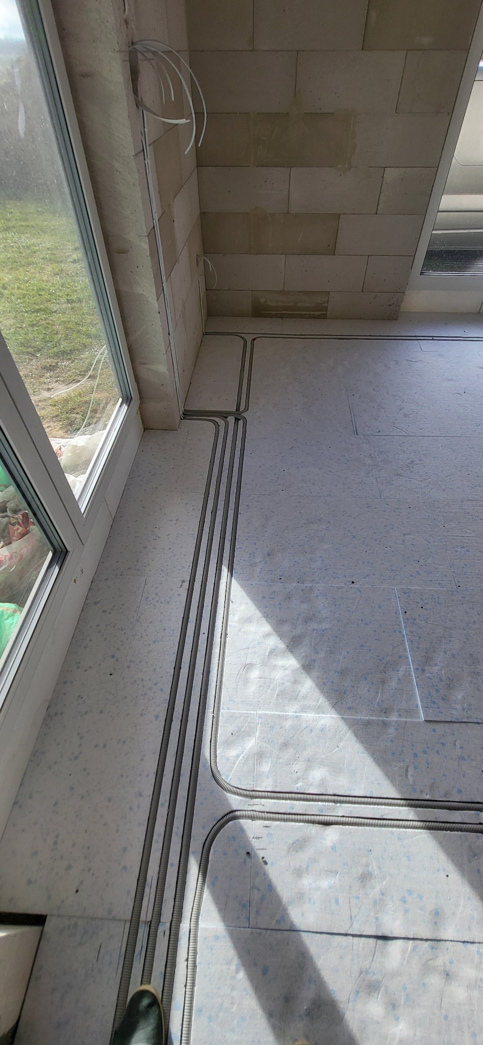 Szare, karbowane rurki ochronne na kable elektryczne ułożone równolegle na podłodze w surowym stanie, prowadzące od okna wzdłuż ściany z betonu komórkowego. Widoczne białe przewody wystające ze...