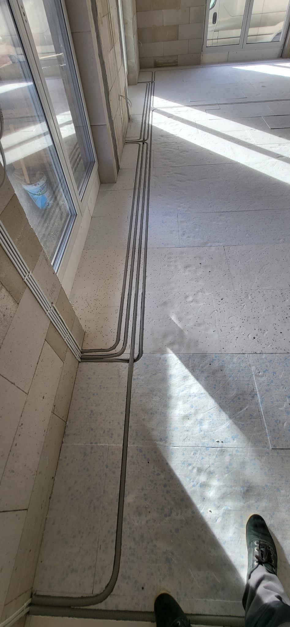 Wnętrze w trakcie prac wykończeniowych: widok z góry na trzy szare peszle elektryczne prowadzone po podłodze wzdłuż ściany z oknem, z fragmentem stopy w czarnym bucie w prawym dolnym rogu.