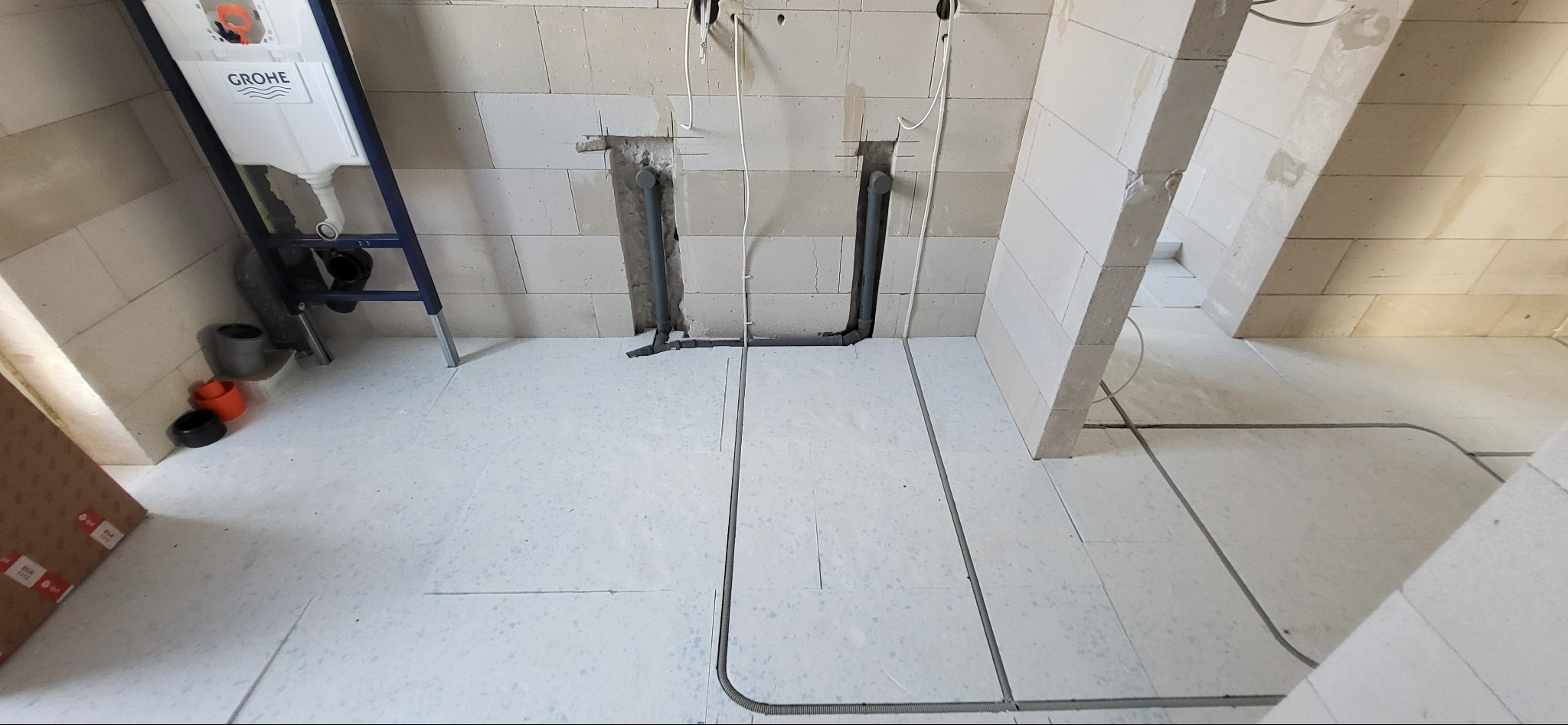 Surowy stan łazienki w trakcie prac: widoczne podłączenia kanalizacyjne, rury w ścianach z betonu komórkowego, oraz rurki instalacji elektrycznej w podłodze, zbiornik WC marki Grohe.