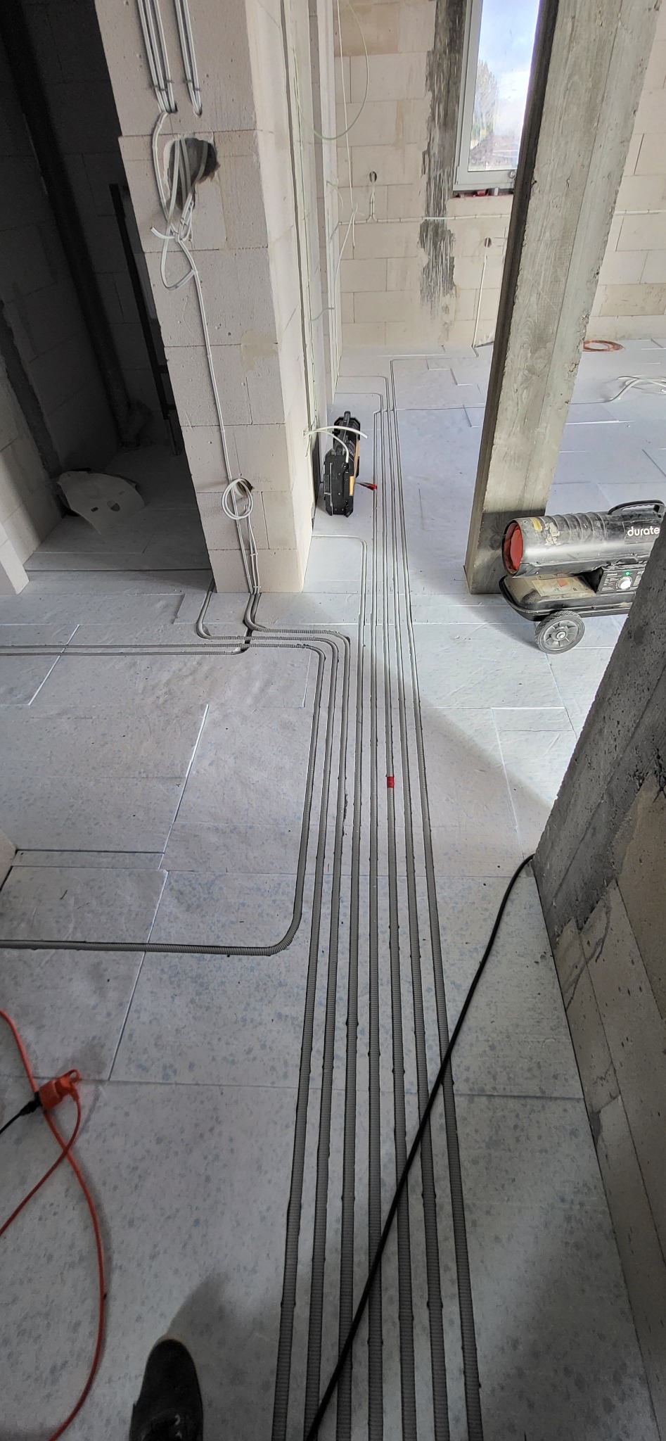 Instalacja elektryczna w surowym stanie: widoczne kable w peszlach ułożone w bruzdach w podłodze, narzędzia elektryczne i nagrzewnica w tle.