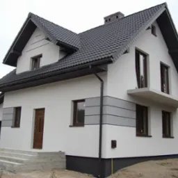 Nowoczesny dom jednorodzinny z ciemną dachówką, białymi ścianami z szarymi akcentami, brązowymi oknami i betonowymi schodami wejściowymi w trakcie budowy.