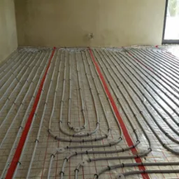 Szare rury systemu ogrzewania podłogowego ułożone równolegle na folii z czerwoną siatką w surowym wnętrzu z oknem.