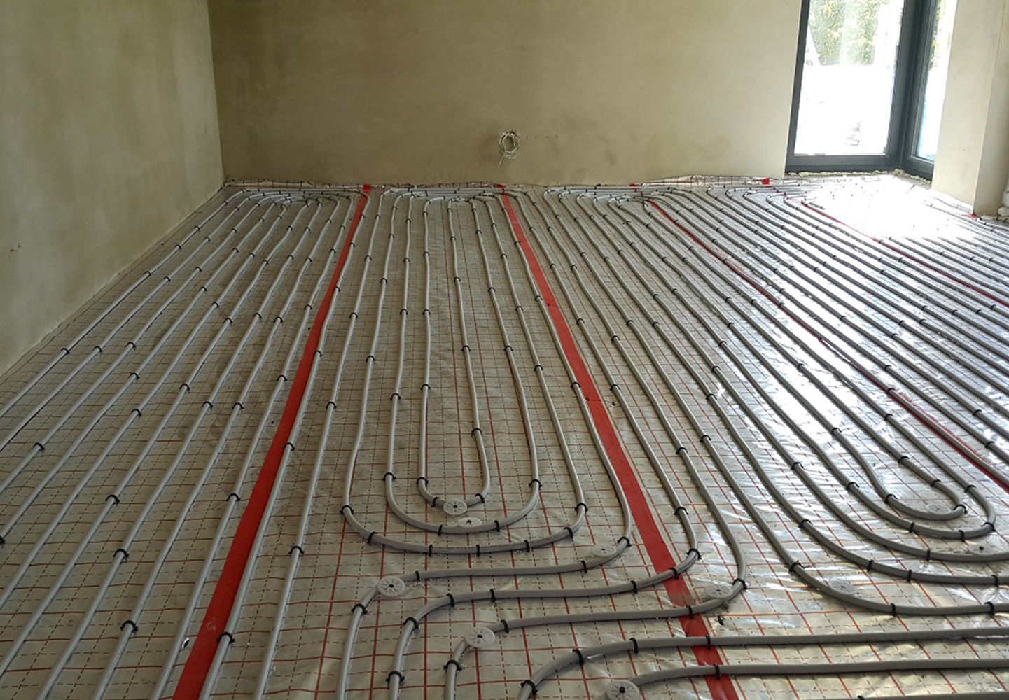 Szare rury systemu ogrzewania podłogowego ułożone równolegle na folii z czerwoną siatką w surowym wnętrzu z oknem.
