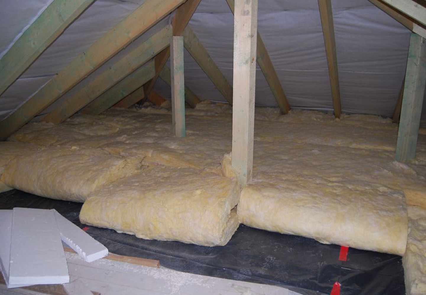 Izolacja poddasza wełną mineralną, widoczne krokwie i czarna folia izolacyjna.