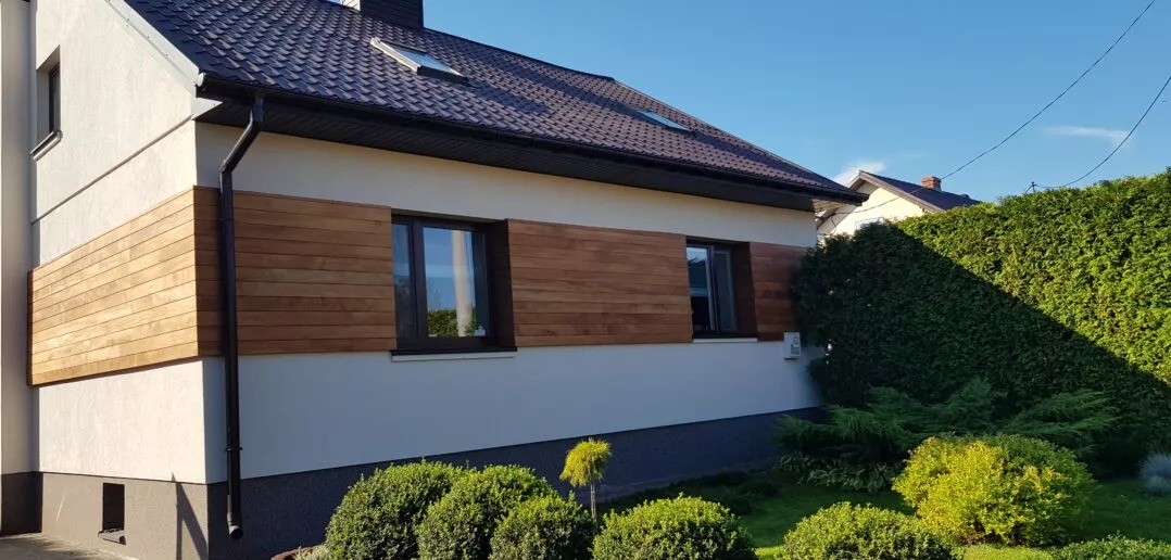 Elewacja domu jednorodzinnego z drewnianą okładziną i ciemnymi oknami, widok z zewnątrz na tle zielonego ogrodu i błękitnego nieba.