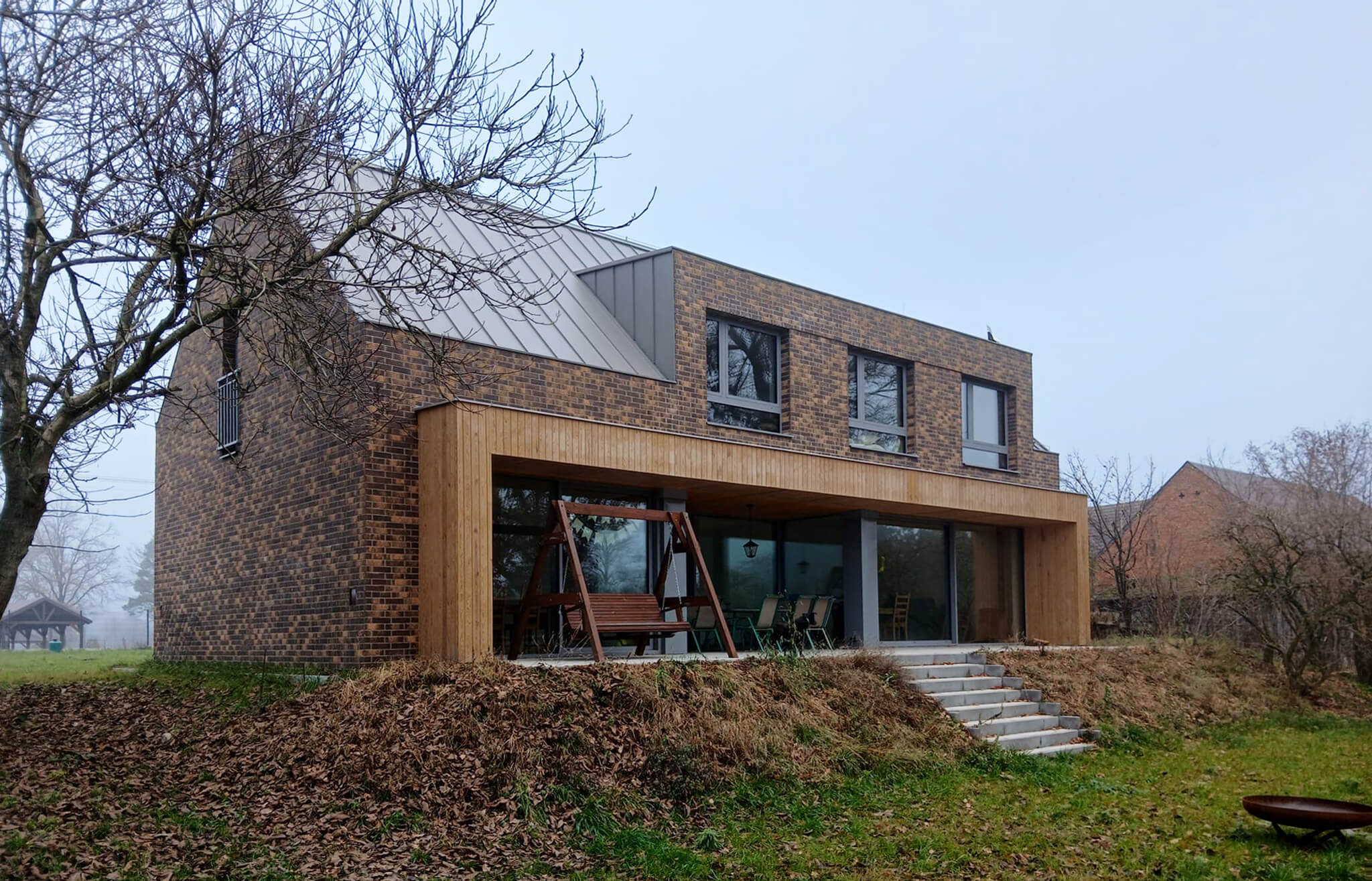 Nowoczesny dom z cegły klinkierowej z dużymi oknami i drewnianą pergolą, widok z ogrodu w pochmurny dzień.