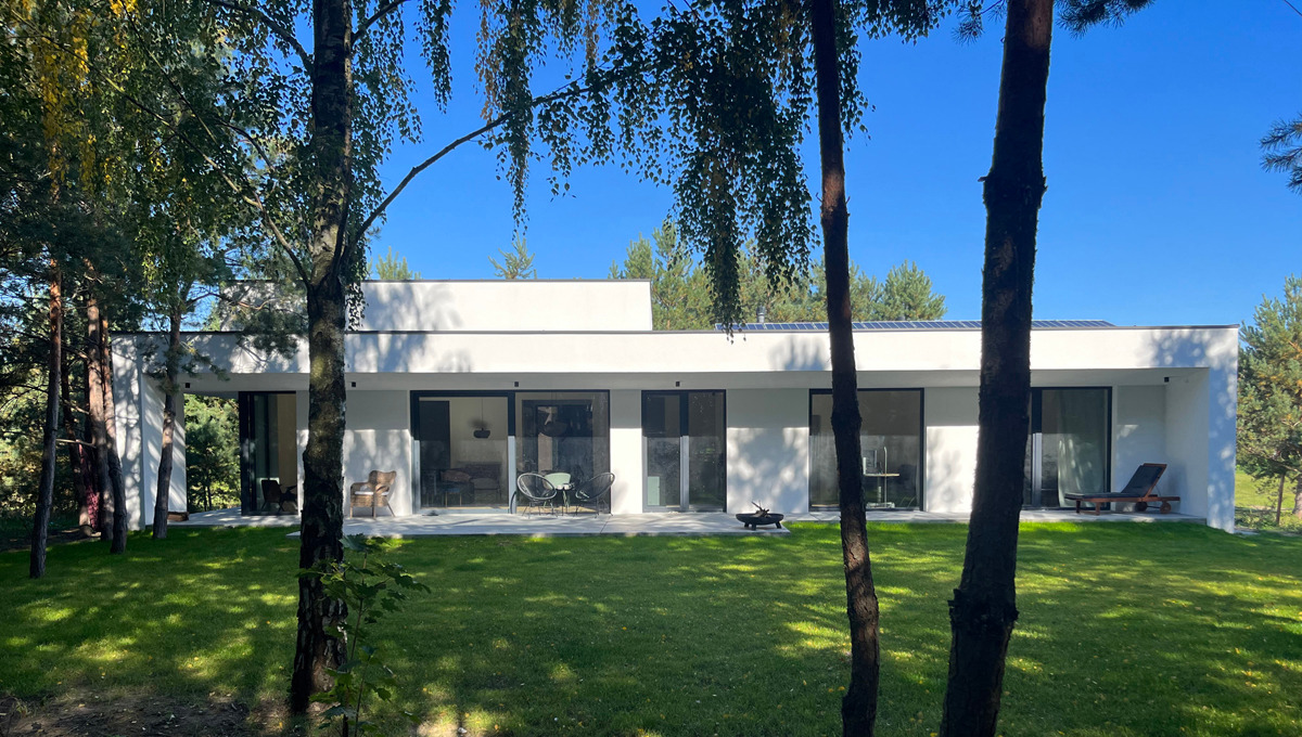 Nowoczesny, parterowy dom z białymi ścianami i dużymi przeszkleniami, widoczny zza drzew na tle błękitnego nieba i zielonego trawnika.