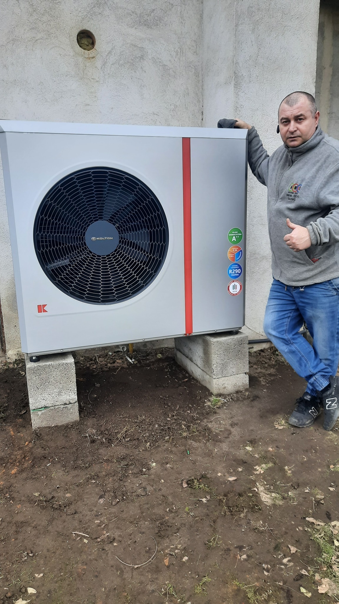 Zewnętrzna jednostka pompy ciepła Kolton zamontowana na betonowych bloczkach, z widocznymi naklejkami informacyjnymi o klasie energetycznej i czynniku chłodniczym R290, obok stoi monter...