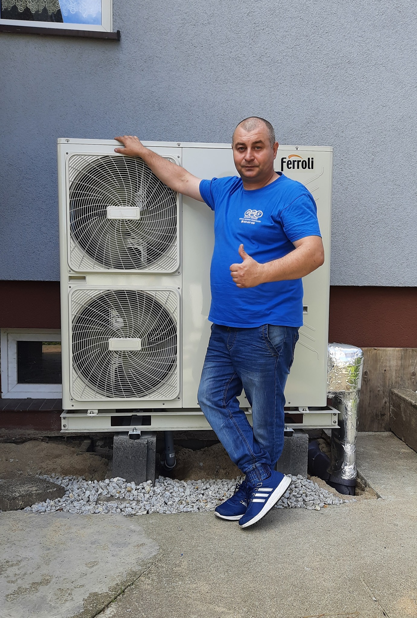 Mężczyzna w niebieskiej koszulce z logo firmy, stojący obok zainstalowanej pompy ciepła marki Ferroli na betonowych blokach i żwirowym podłożu, z uniesionym kciukiem w górę.