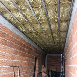Firma ogólnobudowlana - Poddasze w trakcie ocieplania: widoczne metalowe profile sufitowe, pomiędzy którymi umieszczona jest izolacja z wełny mineralnej, ściany z czerwonej cegły, drabina oparta o ścianę, w tle materiały...