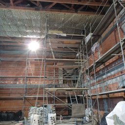 Firma ogólnobudowlana - Poddasze w trakcie izolacji, widoczne rusztowanie, częściowo ocieplony strop wełną mineralną, ceglana ściana w tle i worki z materiałami budowlanymi na podłodze.