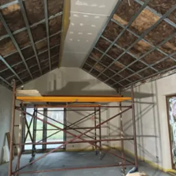 Poddasze w trakcie ocieplania wełną mineralną, widoczny metalowy stelaż i częściowo zamontowana płyta gipsowo-kartonowa, rusztowanie w pomieszczeniu, rękawice robocze na podłodze.