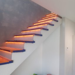 Firma ogólnobudowlana - Schody z podświetleniem LED na stopniach, zabezpieczone niebieską taśmą malarską, ściana w odcieniach szarości i bieli, widoczne materiały budowlane i stołek malarski.