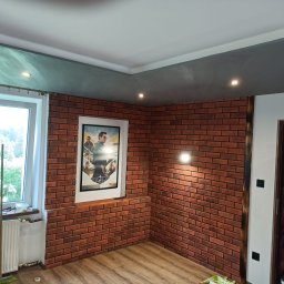 Firma ogólnobudowlana - Pokój z dekoracyjną ścianą z cegły, drewnianą podłogą i plakatem filmu Top Gun w ramie. Widoczne również okno z grzejnikiem, sufit w dwóch odcieniach szarości z oświetleniem punktowym i fragment...