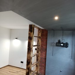 Firma ogólnobudowlana - Wnętrze z drewnianym regałem o opalanej strukturze, ceglaną ścianą i szarą ścianą z ukrytymi instalacjami pod telewizor, parkiet na podłodze, biała ściana z czarnym gniazdkiem.