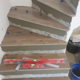 Montaż płytek imitujących drewno na schodach, widoczne poziomice, niebieskie krzyżyki dystansowe i wiadro z zaprawą.