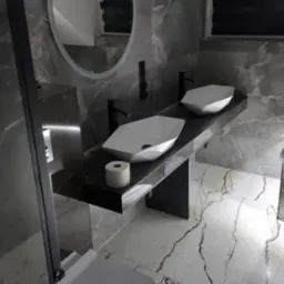 Nowoczesna łazienka z dwiema geometrycznymi umywalkami na czarnym blacie, okrągłe lustro z podświetleniem, marmurowe płytki na ścianach i podłodze z wyrazistym wzorem żyłkowania, wisząca toaleta.