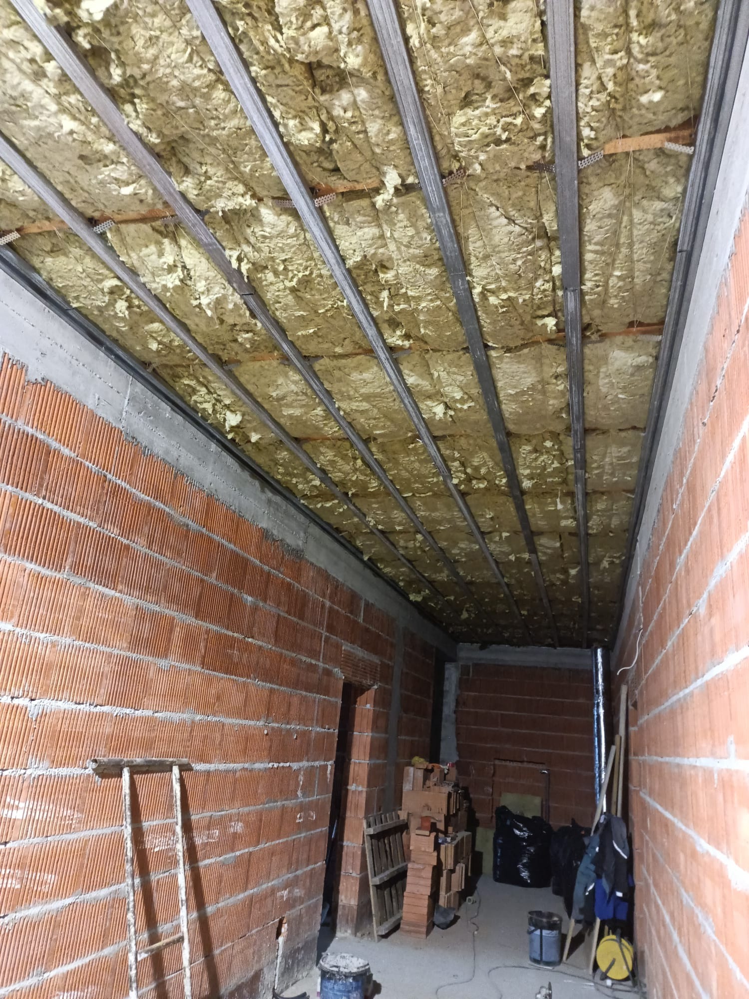 Poddasze w trakcie ocieplania: widoczne metalowe profile sufitowe, pomiędzy którymi umieszczona jest izolacja z wełny mineralnej, ściany z czerwonej cegły, drabina oparta o ścianę, w tle materiały...
