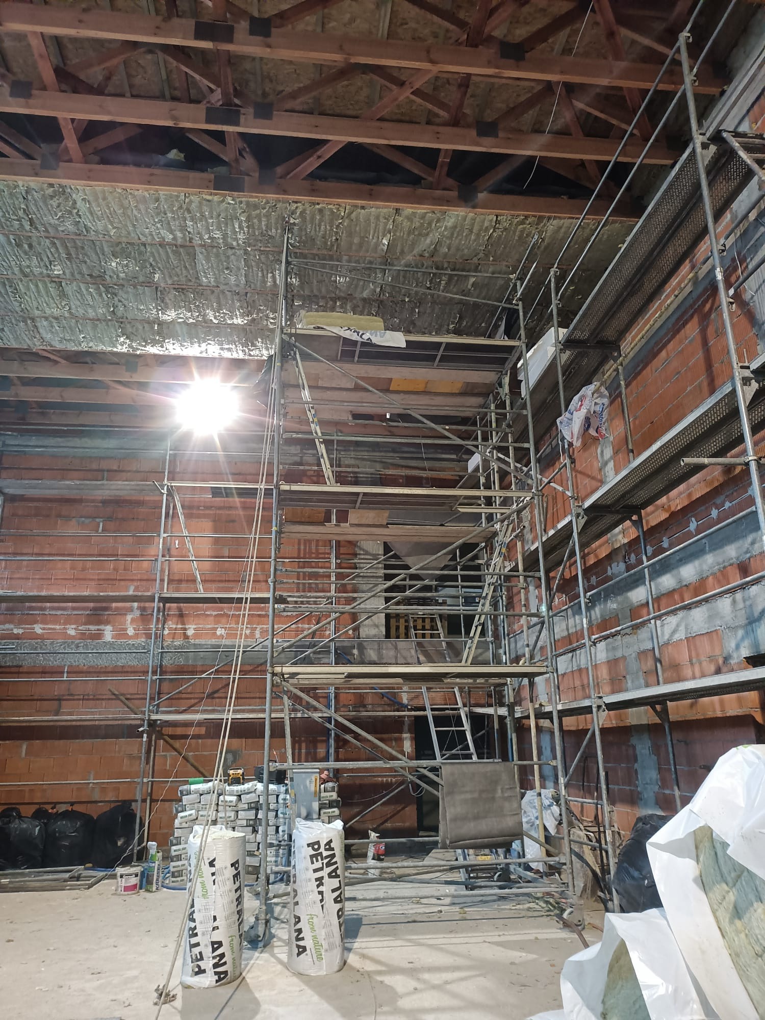 Poddasze w trakcie izolacji, widoczne rusztowanie, częściowo ocieplony strop wełną mineralną, ceglana ściana w tle i worki z materiałami budowlanymi na podłodze.