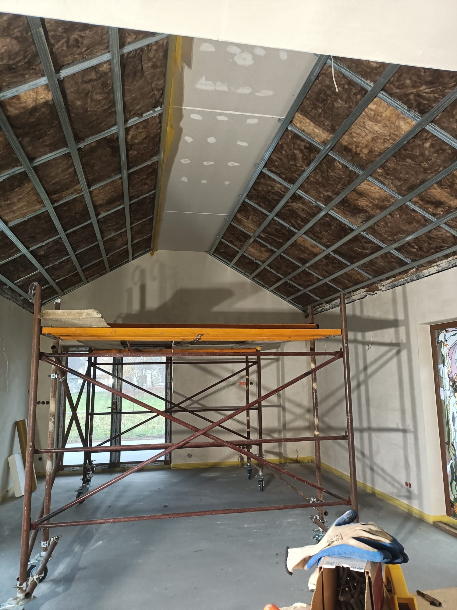 Poddasze w trakcie ocieplania wełną mineralną, widoczny metalowy stelaż i częściowo zamontowana płyta gipsowo-kartonowa, rusztowanie w pomieszczeniu, rękawice robocze na podłodze.