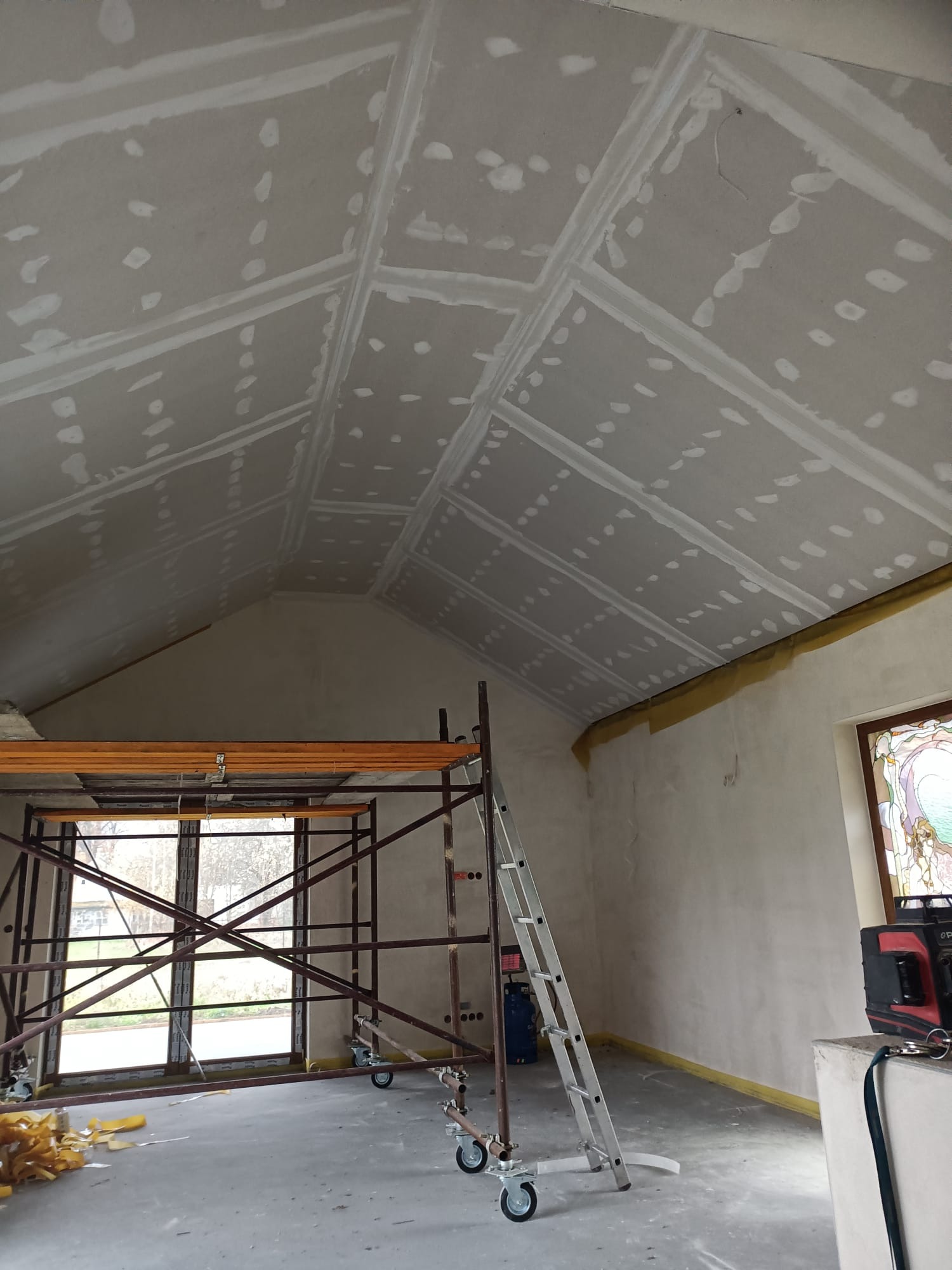 Skośny sufit poddasza z płyt kartonowo-gipsowych, na łączeniach widoczna masa szpachlowa, rusztowanie przy oknie, drabina aluminiowa, zabezpieczona ściana taśmą malarską.