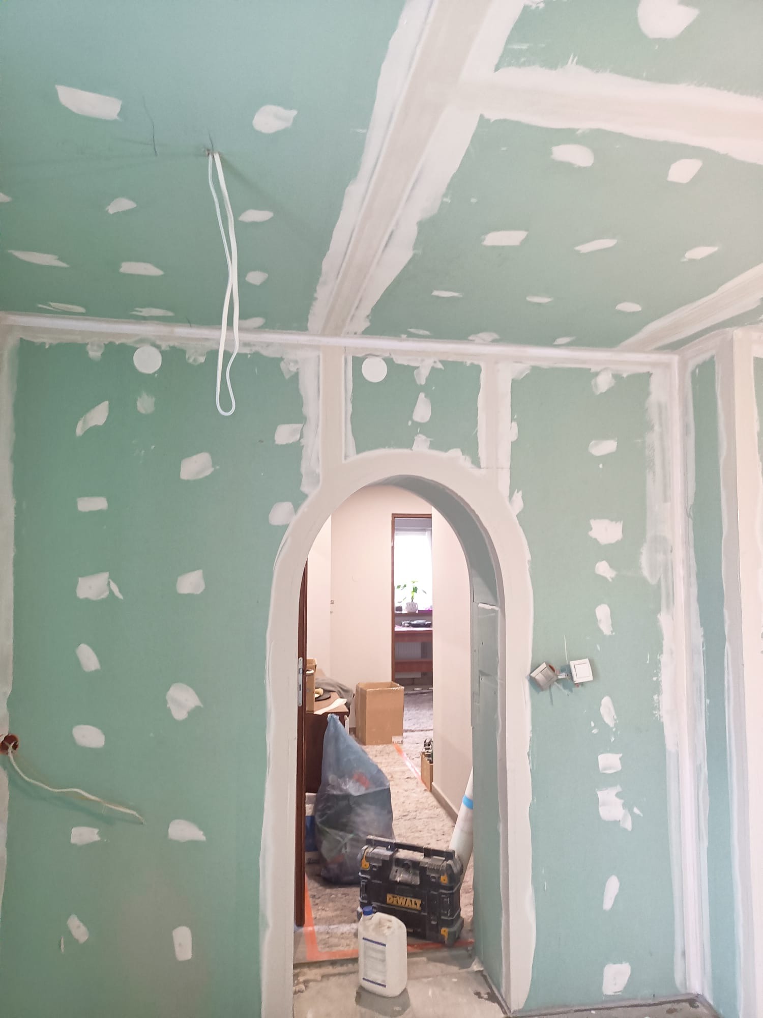 Wnętrze w trakcie montażu płyt kartonowo-gipsowych, widoczne białe fugi maskujące łączenia, surowe ściany w kolorze zielonym, narzędzia DeWalt i worki na podłodze, przejście z łukiem do kolejnego...