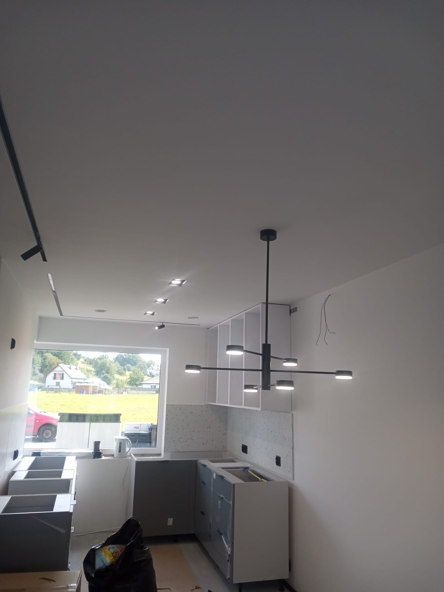 Wnętrze kuchni w trakcie montażu: widoczne szafki kuchenne bez frontów, nowoczesna lampa sufitowa, listwa oświetleniowa, okno z widokiem na zewnątrz, ściany przygotowane do malowania i częściowo...