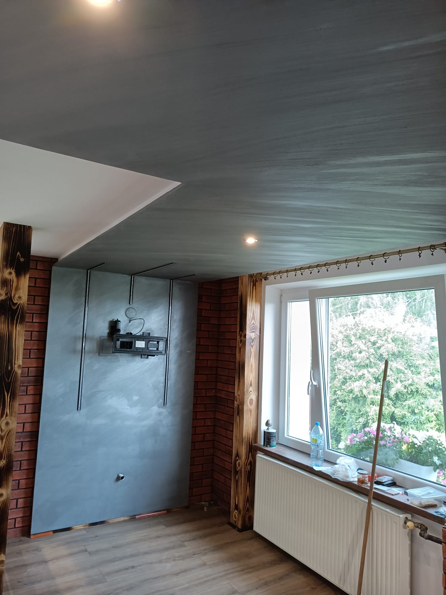 Wnętrze pokoju po remoncie: ściana z cegły klinkierowej, drewniane belki, szara ściana z zamontowanym uchwytem na telewizor, szary sufit, okno z widokiem na zieleń.