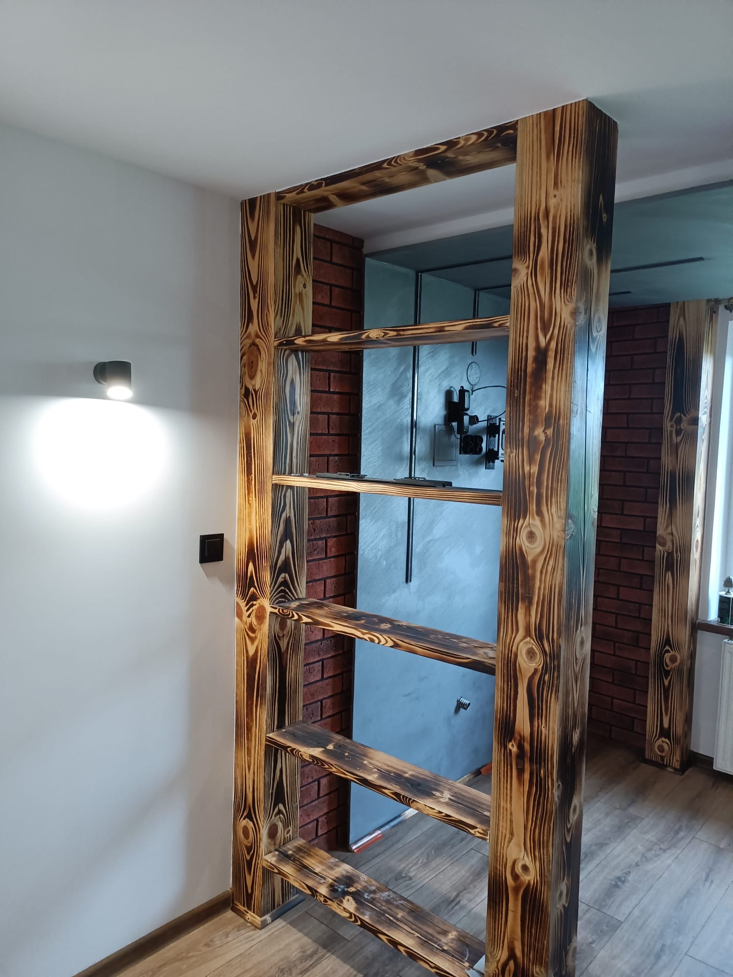 Ażurowa ścianka działowa z opalanych desek, tworząca regał w pokoju z ceglaną ścianą i szarą ścianą w tle.