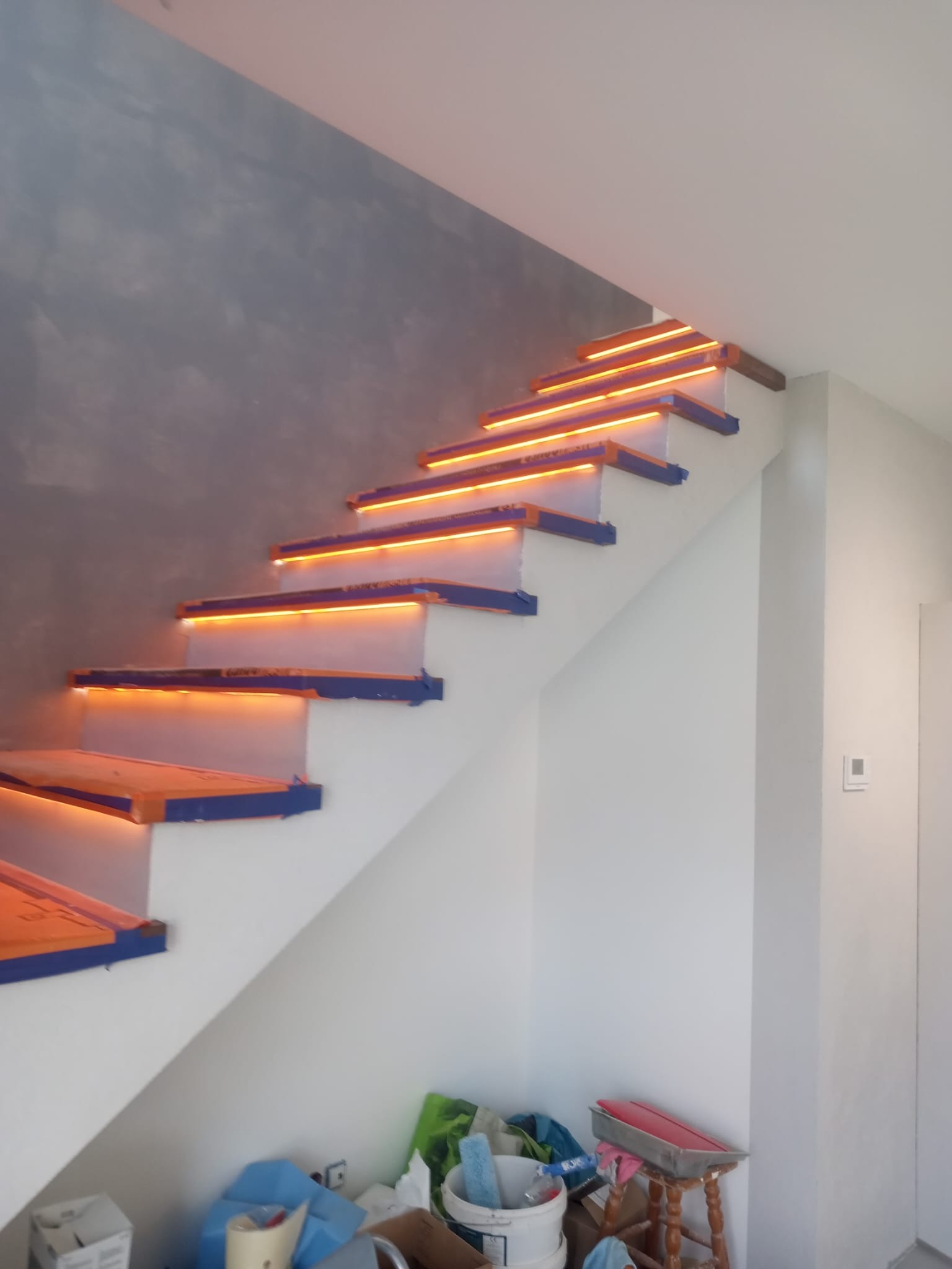 Schody z podświetleniem LED na stopniach, zabezpieczone niebieską taśmą malarską, ściana w odcieniach szarości i bieli, widoczne materiały budowlane i stołek malarski.
