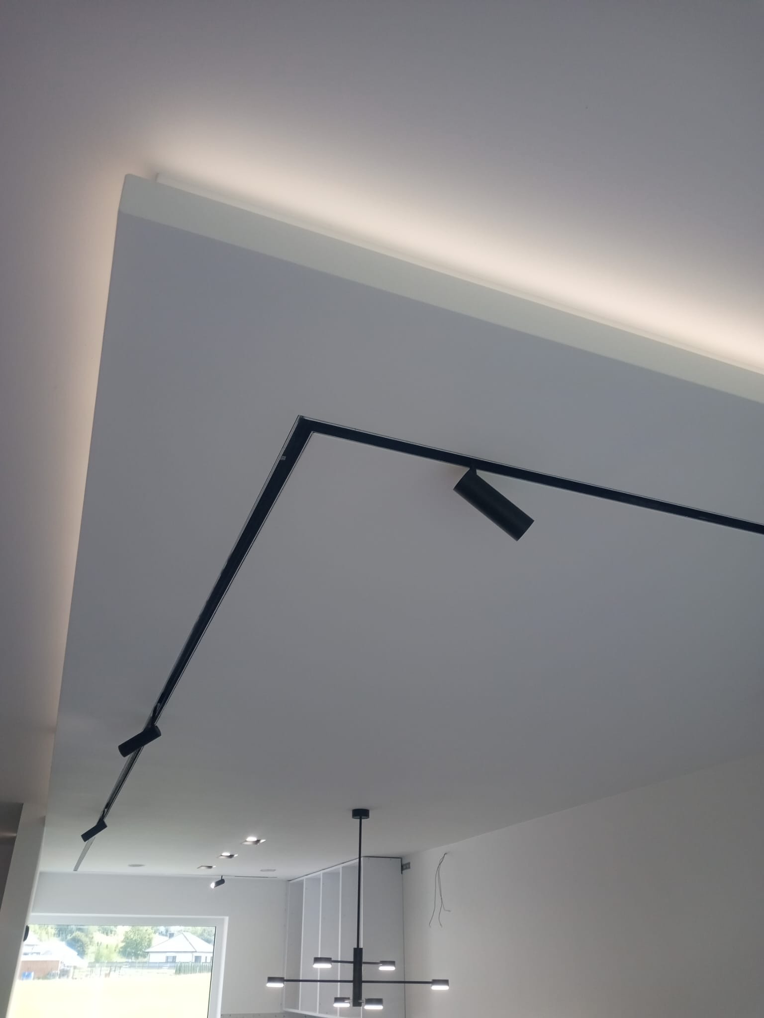 Nowoczesne oświetlenie sufitowe z czarnymi reflektorami punktowymi na szynoprzewodzie, ukryte oświetlenie LED w listwach, widok z dołu na biały sufit i fragment wnętrza z oknem.