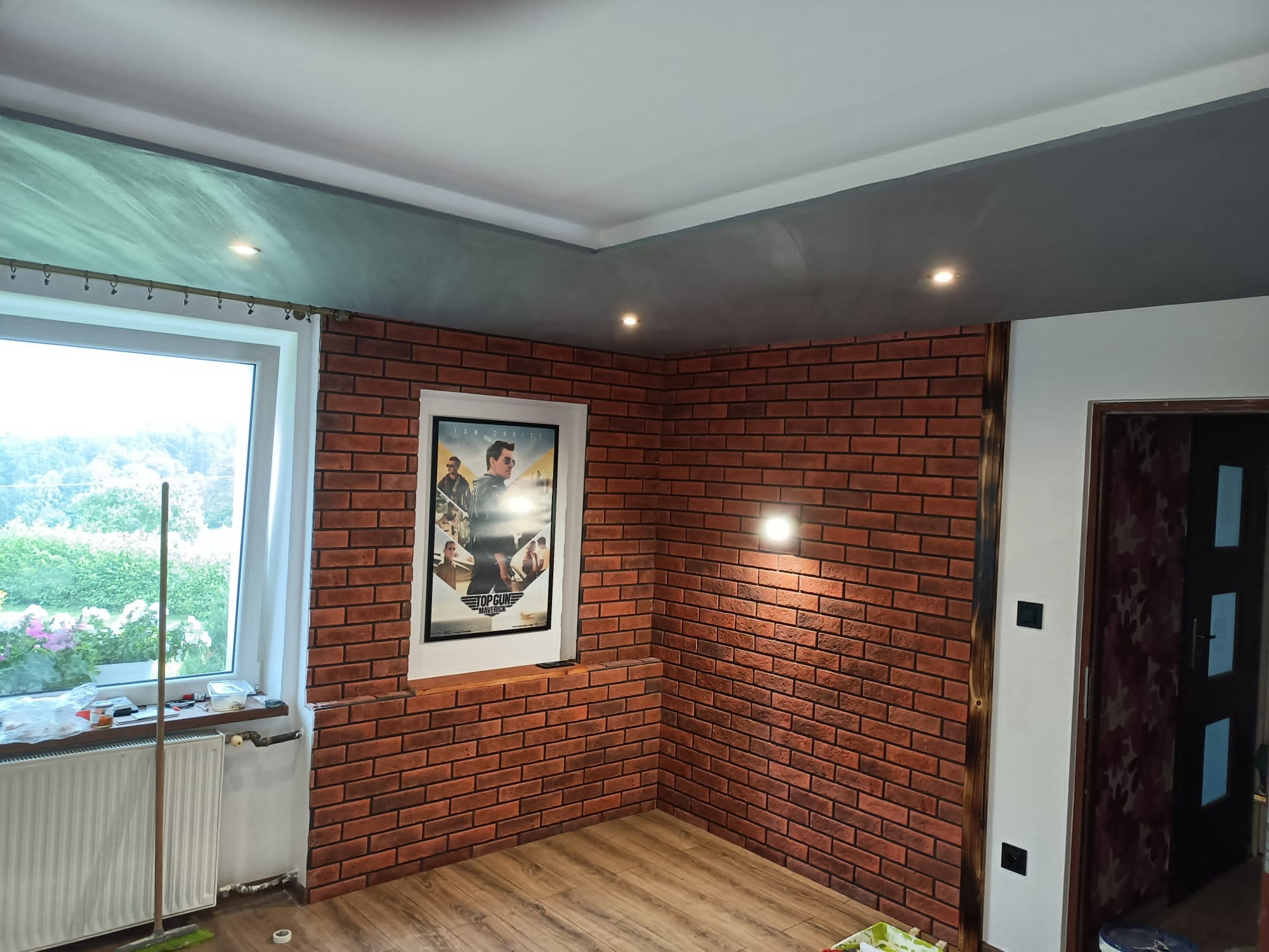 Pokój z dekoracyjną ścianą z cegły, drewnianą podłogą i plakatem filmu Top Gun w ramie. Widoczne również okno z grzejnikiem, sufit w dwóch odcieniach szarości z oświetleniem punktowym i fragment...