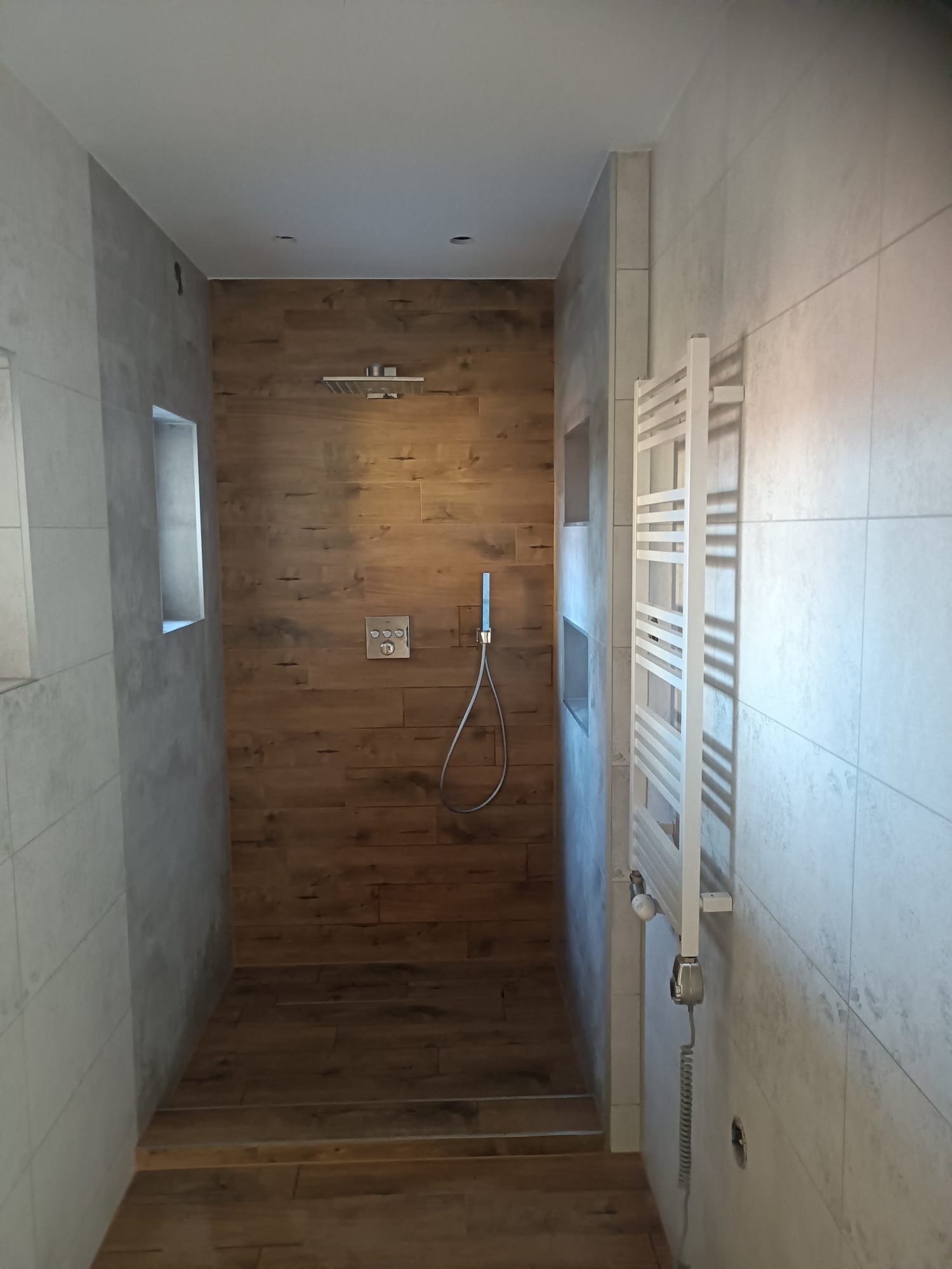 Nowoczesna kabina prysznicowa typu walk-in z drewnopodobnymi płytkami na ścianie i podłodze, wyposażona w deszczownicę, termostat i grzejnik drabinkowy w kolorze białym, z wnękami w ścianach...