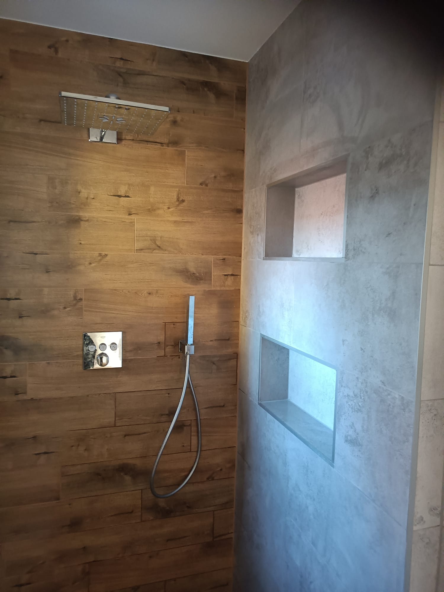 Nowoczesna kabina prysznicowa z drewnopodobnymi płytkami na jednej ścianie i szarą, betonową fakturą na drugiej, z wnękami na kosmetyki.