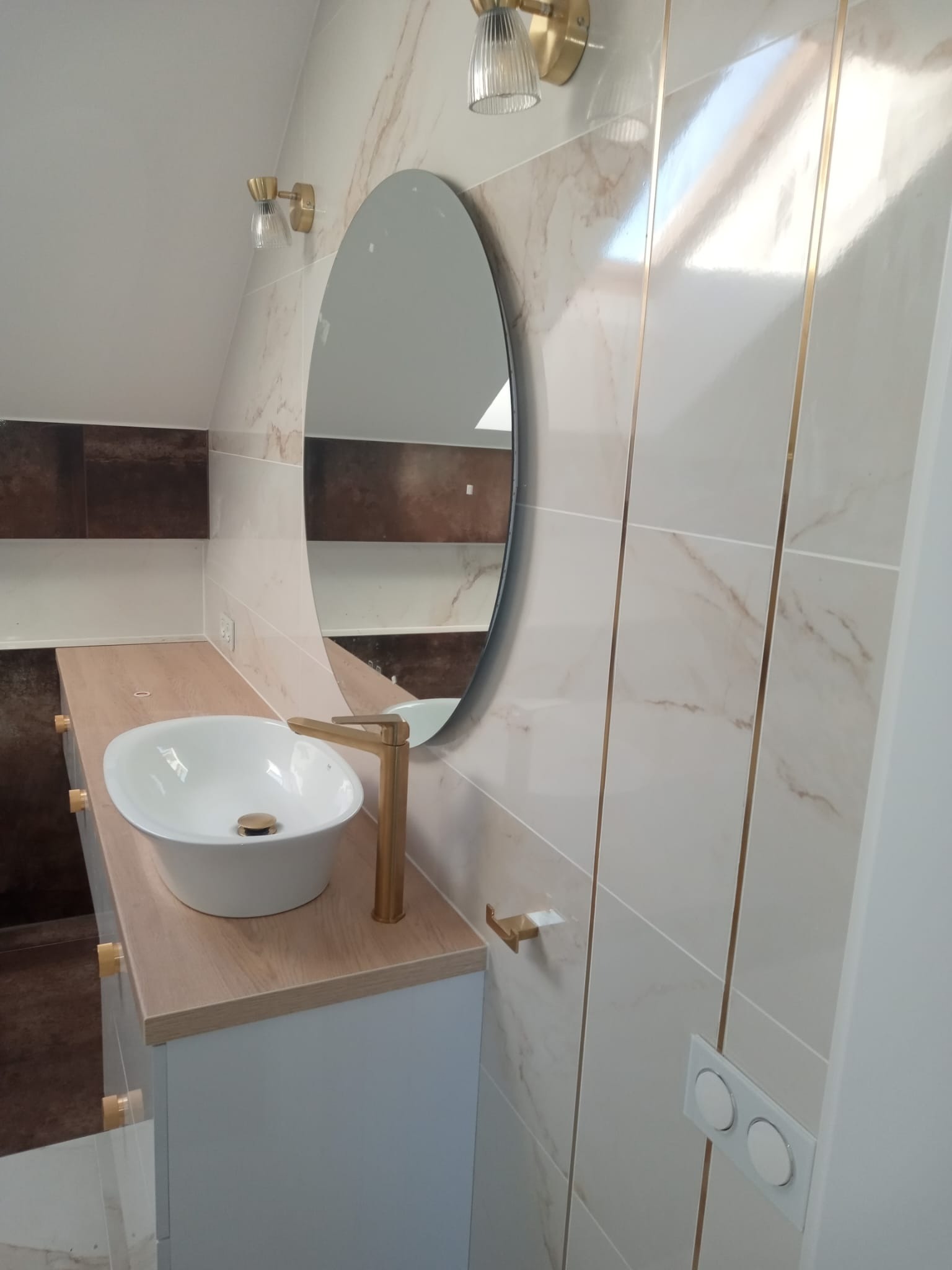 Nowoczesna łazienka na poddaszu z owalnym lustrem, białą umywalką nablatową z złotą armaturą, płytkami imitującymi marmur z delikatnymi złotymi listwami oraz oświetleniem w stylu glamour.