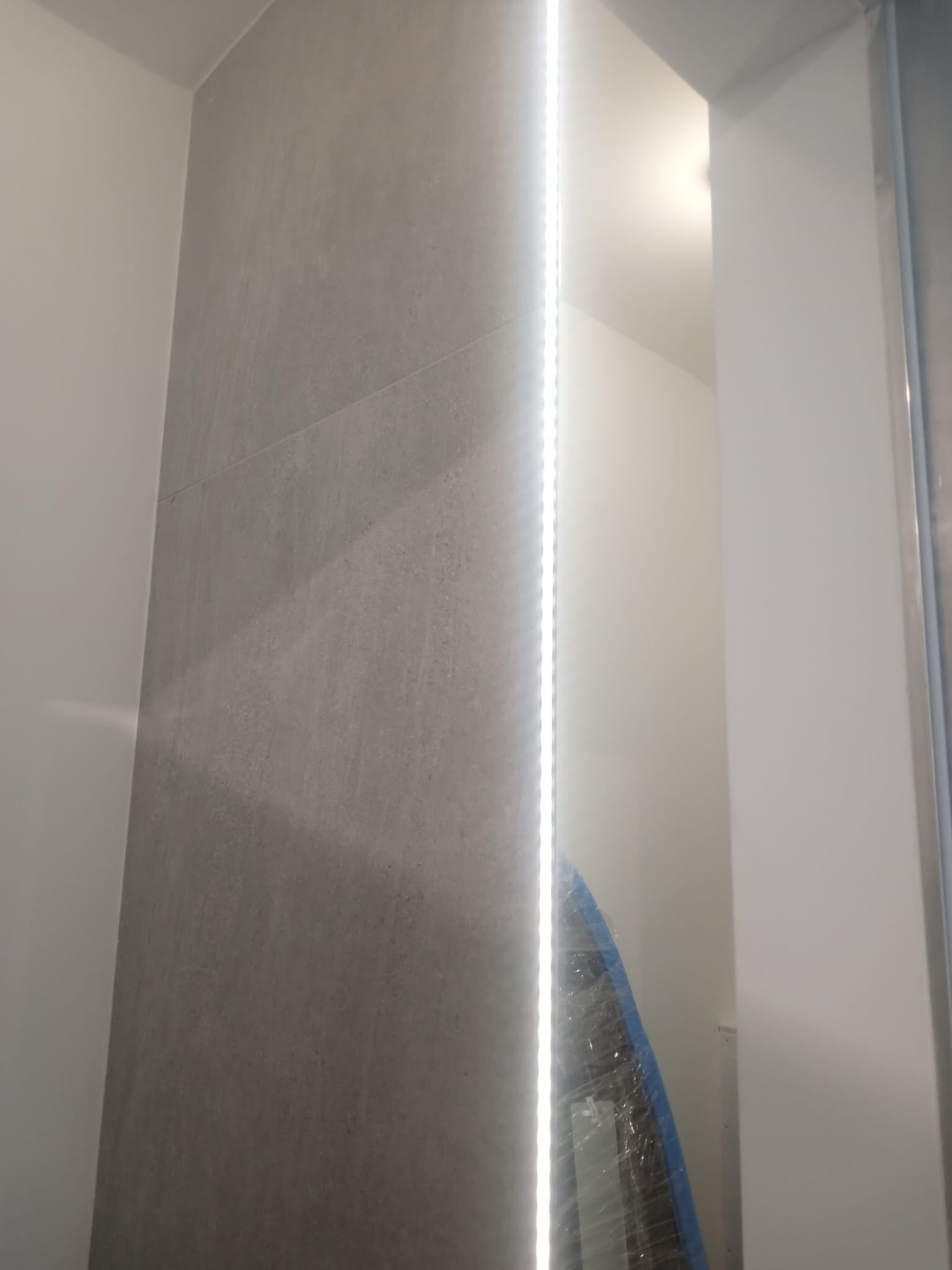 Szara ściana wyłożona płytkami z pionowym oświetleniem LED, widoczny fragment zabezpieczonego folią elementu wyposażenia.